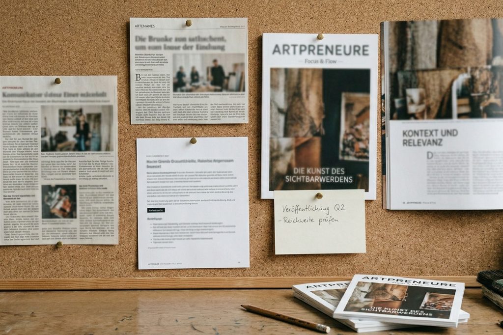 Ein dokumentarisches Foto einer gut gepinnten Korkwand in einem professionellen K&uuml;nstlerstudio. Vier verschiedene, leicht unscharfe Presseartikel, Clippings und Ausdrucke sind sauber mit Messingrei&szlig;zwecken befestigt. Sie zeigen unterschiedliche Layouts und Textbl&ouml;cke. Ein offenes 'Artpreneure'-Magazin und ein Post-it-Notiz mit handschriftlichem Text sind subtil sichtbar. Das warme, nat&uuml;rliche Licht betont die Texturen der Korkwand und der Papierbelege. Der Text im Bild ist maximal subtil gehalten.
